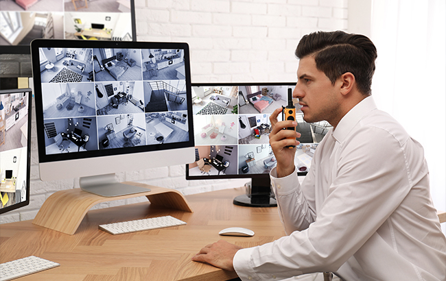 Security professional monitoring multiple surveillance camera feeds on computer screens in a commercial security control room.