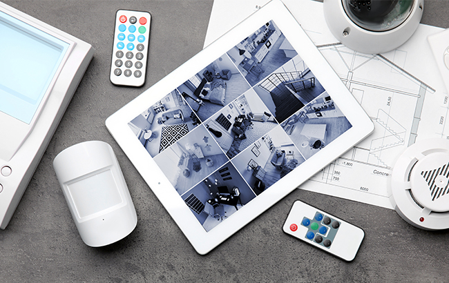 Tablet displaying multiple home security camera feeds with motion sensors and alarm devices on a table.